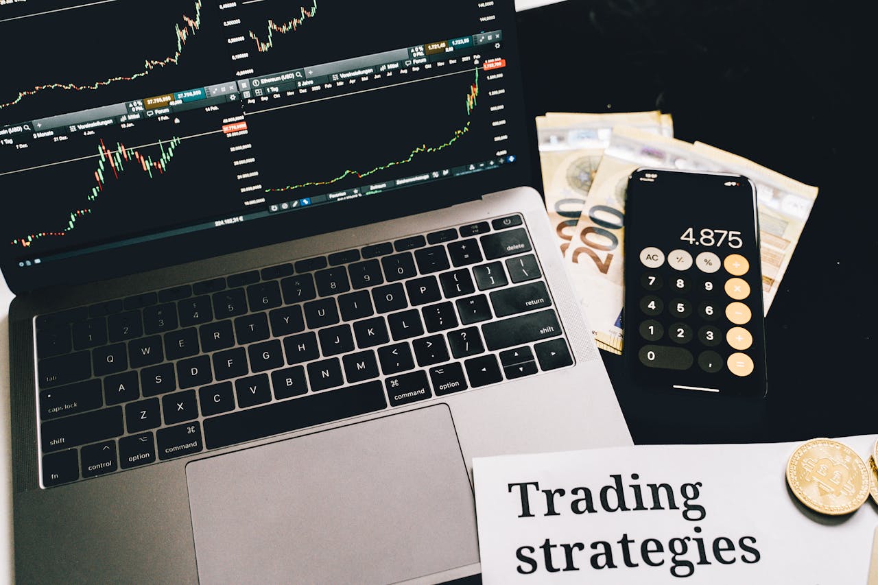 A laptop showing stock market graphs with a smartphone calculator, euro bills, coins, and trading strategies paper.