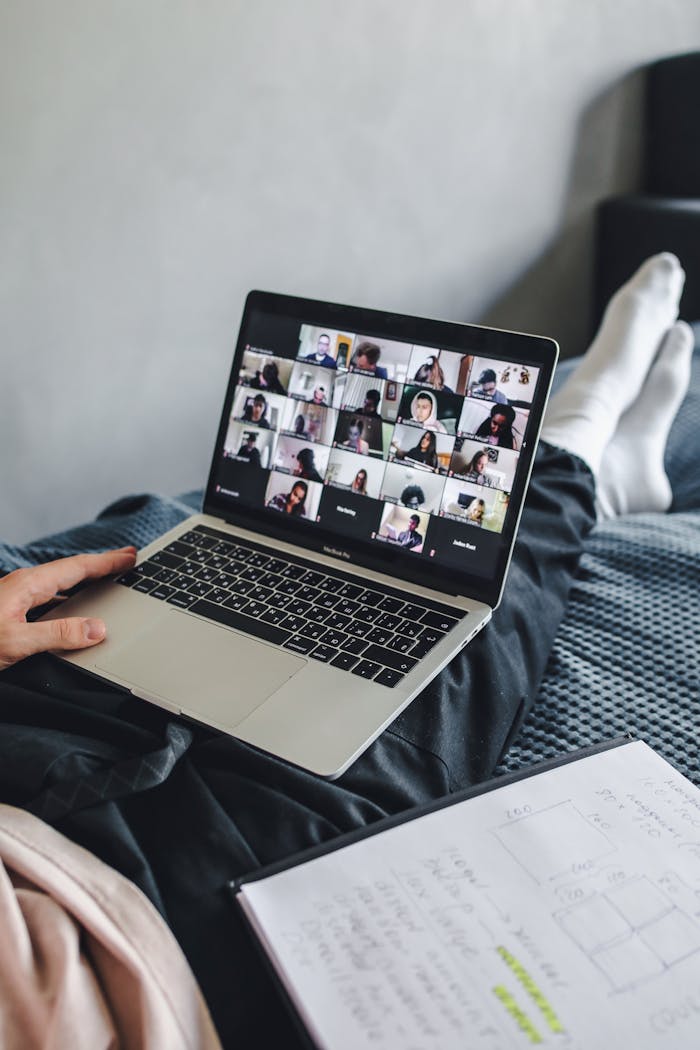 Adult sitting on bed using laptop for video conference and note-taking.